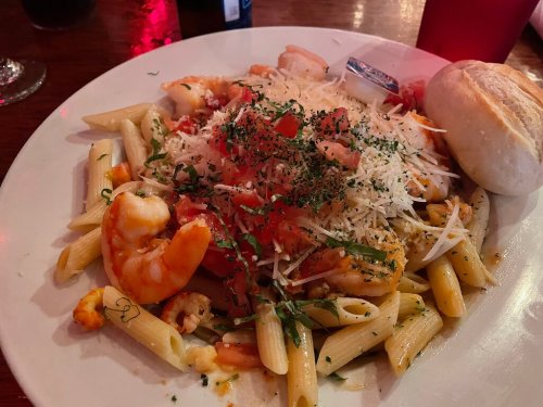 A plate of pasta with shrimp, topped with cheese and herbs, served with a bread roll on the side.