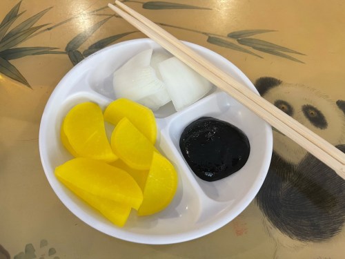 A white dish with yellow pickled vegetables, sliced onions, and a dark sauce, accompanied by chopsticks.