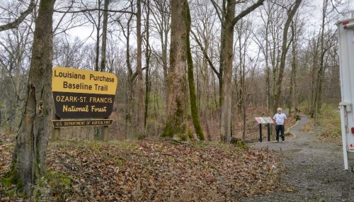 A person stands near a sign for the Louisiana Purchase Baseline Trail in a wooded area of Ozark-St. Francis National Forest.