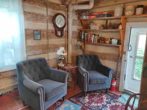 Cozy interior with two gray armchairs, a small table, a clock, and a bookshelf against a rustic wooden wall.