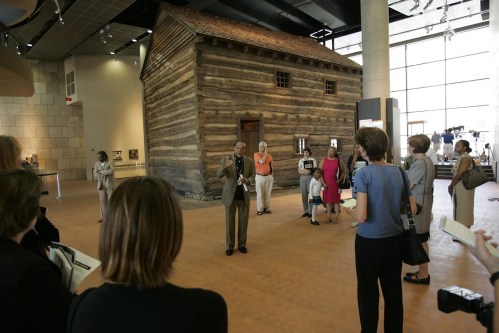 A group of people gathers around a speaker in front of a wooden cabin exhibit in a museum.