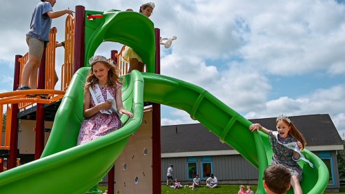 Children play on a colorful slide at a playground, with some wearing crowns and sashes, enjoying a sunny day.