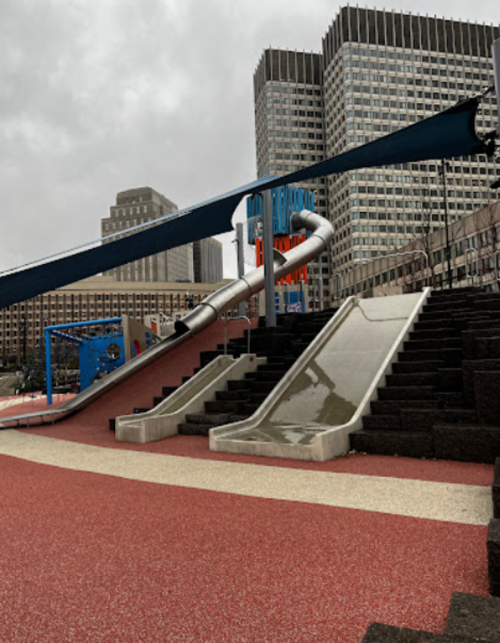 A modern playground featuring a large slide, steps, and colorful rubber flooring, set against a city skyline.