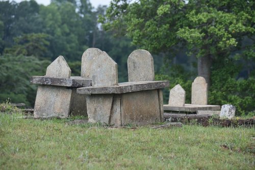 A weathered graveyard with several old tombstones set on a grassy hillside, surrounded by trees.