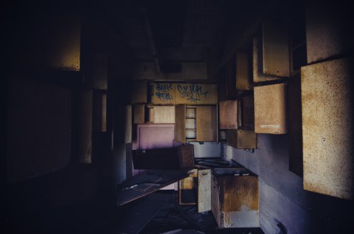 Abandoned room with dark walls, scattered debris, and rusty cabinets, illuminated by dim light.