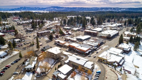 Aerial view of a snowy town with shops, trees, and mountains in the background under a cloudy sky.