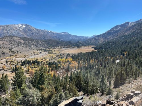 A scenic view of a valley surrounded by mountains, with autumn foliage and a clear blue sky.