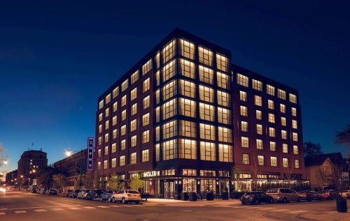 Modern hotel building at night, featuring large windows and illuminated signage, with cars parked along the street.