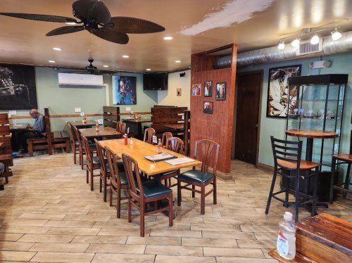A cozy restaurant interior with wooden tables, chairs, and a fan, featuring artwork on the walls and a glass display case.