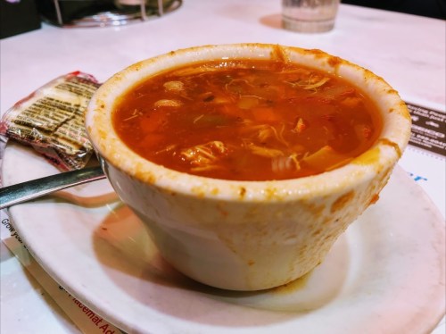 A bowl of hearty soup with chunks of meat and vegetables, served on a white plate with a spoon beside it.