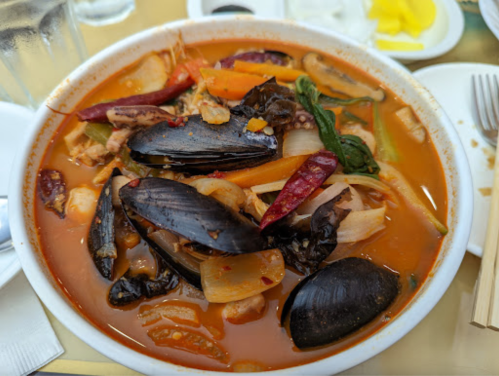 A bowl of spicy seafood soup with mussels, vegetables, and herbs, served on a gold table.