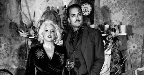A glamorous couple poses in vintage attire against a decorative backdrop, holding a drink and exuding a classic vibe.