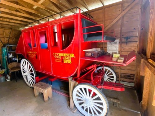 A vintage red stagecoach from 1858, displayed inside a wooden barn with various items around it.