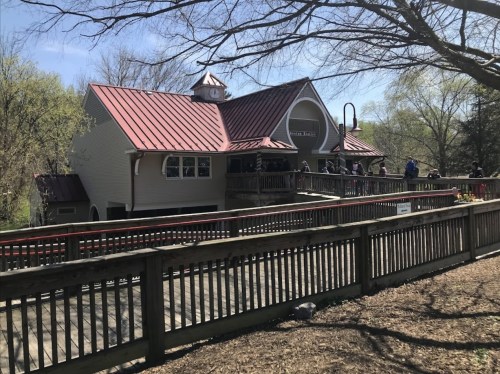 A wooden deck leads to a building with a red roof, surrounded by trees and people enjoying a sunny day.