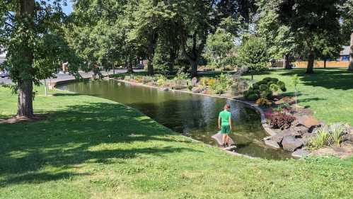 A person in a green shirt walks along a curved pond surrounded by trees and colorful flowers in a park.