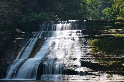 A serene waterfall cascading over rocky ledges, surrounded by lush greenery and dappled sunlight.