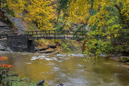 A wooden bridge spans a flowing river, surrounded by vibrant autumn foliage and rocky terrain.