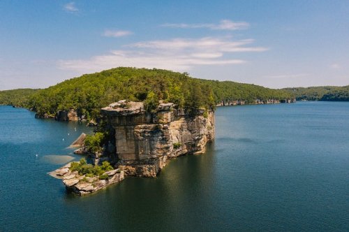 Aerial view of a rocky island surrounded by calm blue water and lush green trees under a clear sky.
