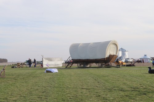 A covered wagon on a grassy field, with people and farm equipment in the background under a cloudy sky.