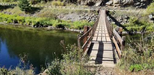 A wooden suspension bridge spans a calm river, surrounded by lush greenery and rocky terrain.