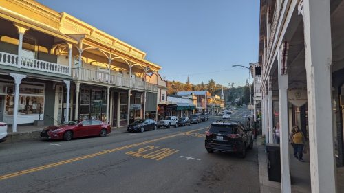 A quiet street lined with historic buildings, shops, and parked cars under a clear blue sky.