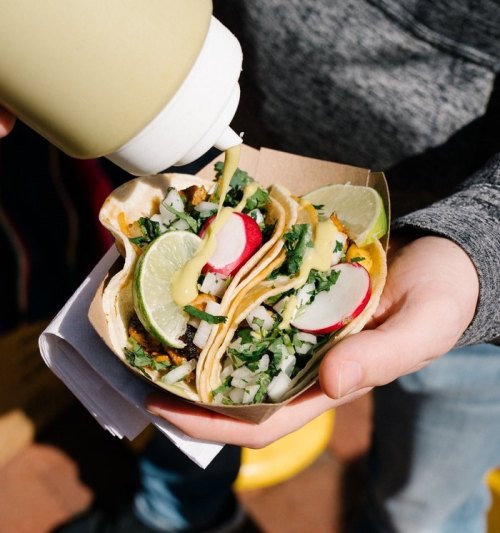 A person holds a tray with two tacos topped with radishes, lime, and green sauce, ready to be enjoyed.