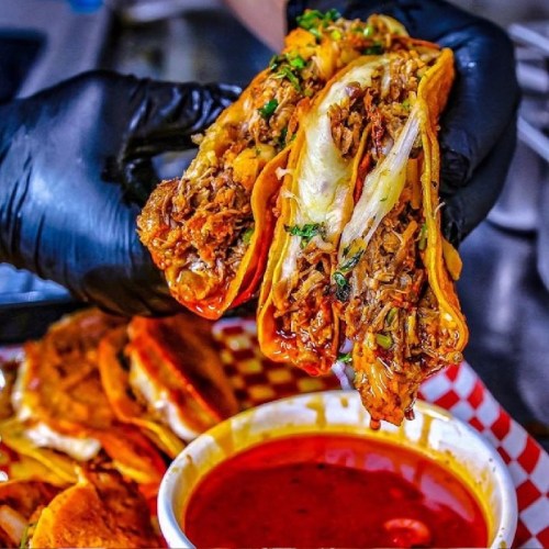 A person wearing black gloves holds two loaded tacos with meat, cheese, and onions, alongside a bowl of red sauce.