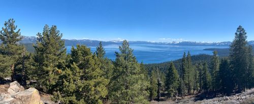 Panoramic view of a lake surrounded by mountains and lush green trees under a clear blue sky.