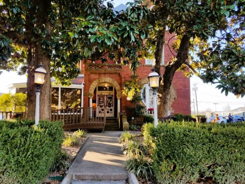 A charming brick building with a welcoming entrance, surrounded by greenery and trees, in a sunny setting.