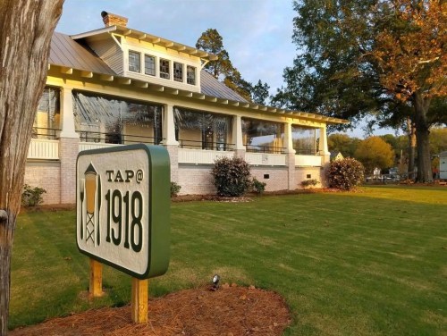 A building with a sign reading "TAP@1918" surrounded by green grass and trees in a sunny setting.