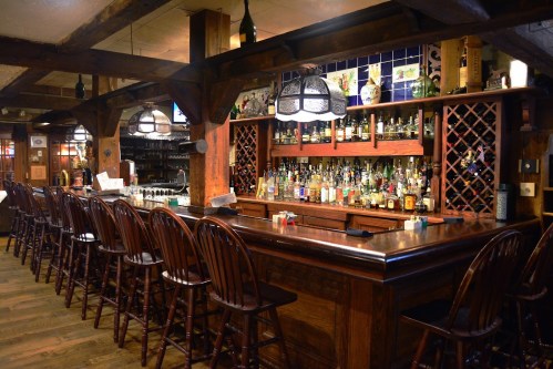 A rustic bar with wooden stools, shelves lined with bottles, and warm lighting in a cozy setting.