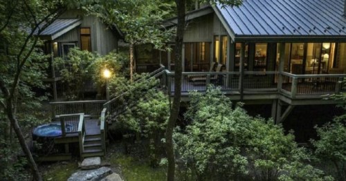 A modern cabin nestled in a lush forest, featuring a deck and hot tub, illuminated by soft outdoor lighting.