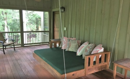 A cozy porch with a green swing bed and decorative pillows, surrounded by wooden walls and large windows.