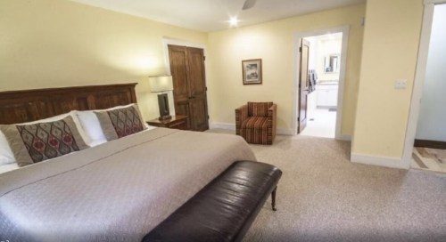 Cozy bedroom with a large bed, a striped chair, and an open door leading to a bathroom. Soft yellow walls.