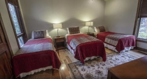 Cozy bedroom with three beds, red bedspreads, lamps, and wooden floors, surrounded by windows.