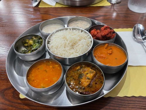 A stainless steel platter with rice, various curries, and side dishes, arranged in small bowls on a wooden table.