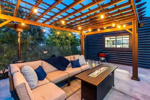 Cozy outdoor patio with a sofa, fire table, and string lights under a wooden pergola, next to a grill and greenery.