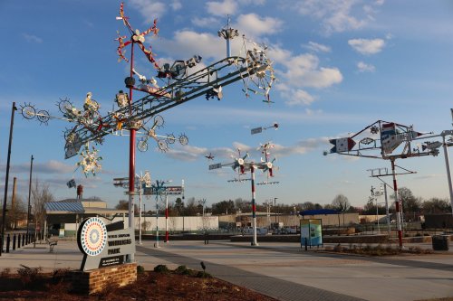 Colorful metal sculptures and wind spinners in a park, with a blue sky and clouds in the background.