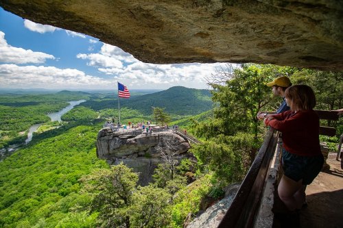 Two people stand on a balcony overlooking a lush green landscape with a river and a flag flying on a rocky outcrop.
