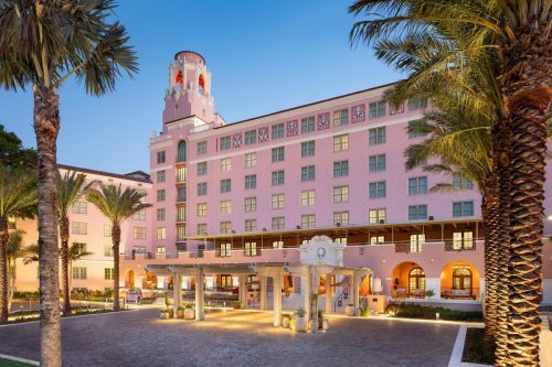 A pink hotel with a tower, surrounded by palm trees, at dusk with lights glowing in the windows.