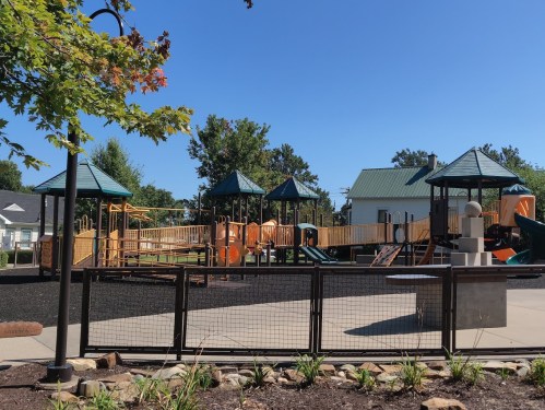 A playground with multiple structures, slides, and climbing areas, surrounded by trees and a clear blue sky.