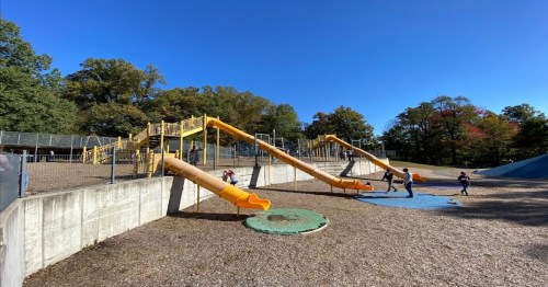 Two large yellow slides at a playground with children playing on a sunny day, surrounded by trees.