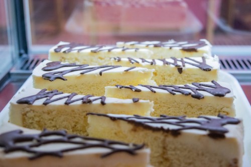 Slices of yellow cake with white frosting and drizzled chocolate on top, displayed in a glass case.