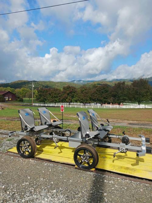 A four-seat rail cart on a yellow platform, set against a backdrop of green hills and a blue sky with clouds.
