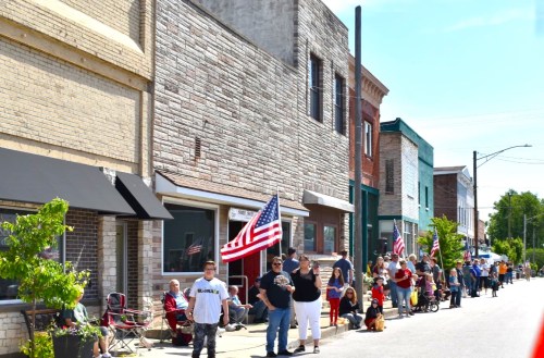 A crowd of people with American flags stands along a street lined with colorful buildings on a sunny day.