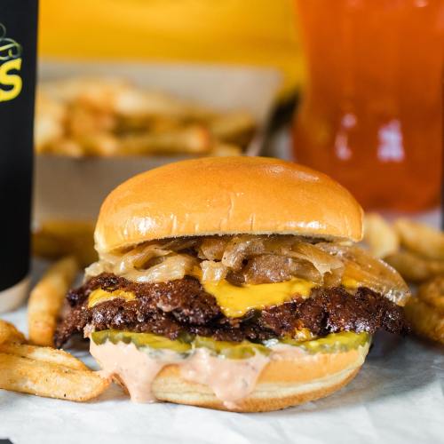 A juicy cheeseburger with melted cheese and grilled onions, served with crispy fries and a drink in the background.