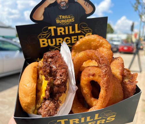 A box of Trill Burgers featuring a cheeseburger and crispy onion rings against a sunny outdoor backdrop.