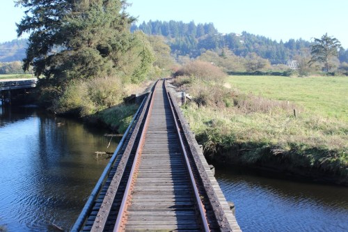A wooden railway bridge stretches over a calm waterway, surrounded by lush greenery and distant hills.