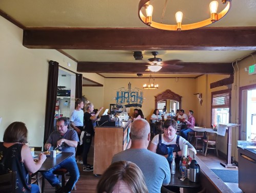 A cozy restaurant interior with diners at tables, staff serving, and a mural on the wall reading "Hash."
