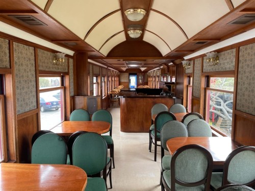 Interior of a vintage train car featuring wooden paneling, green chairs, and tables, with large windows and a bar area.